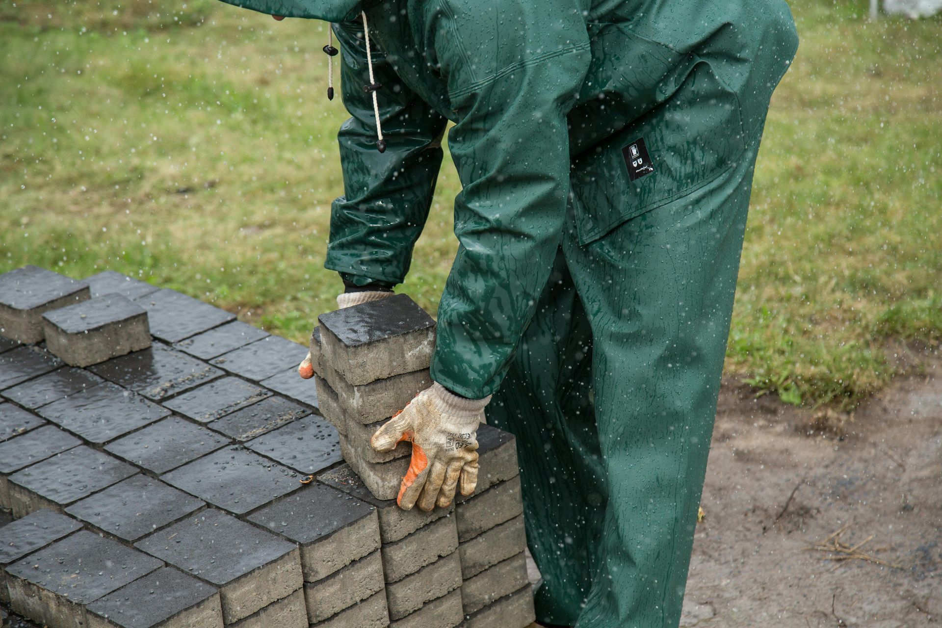 Worker in green rain gear placing roof tiles on a wet shingled roof edge.
