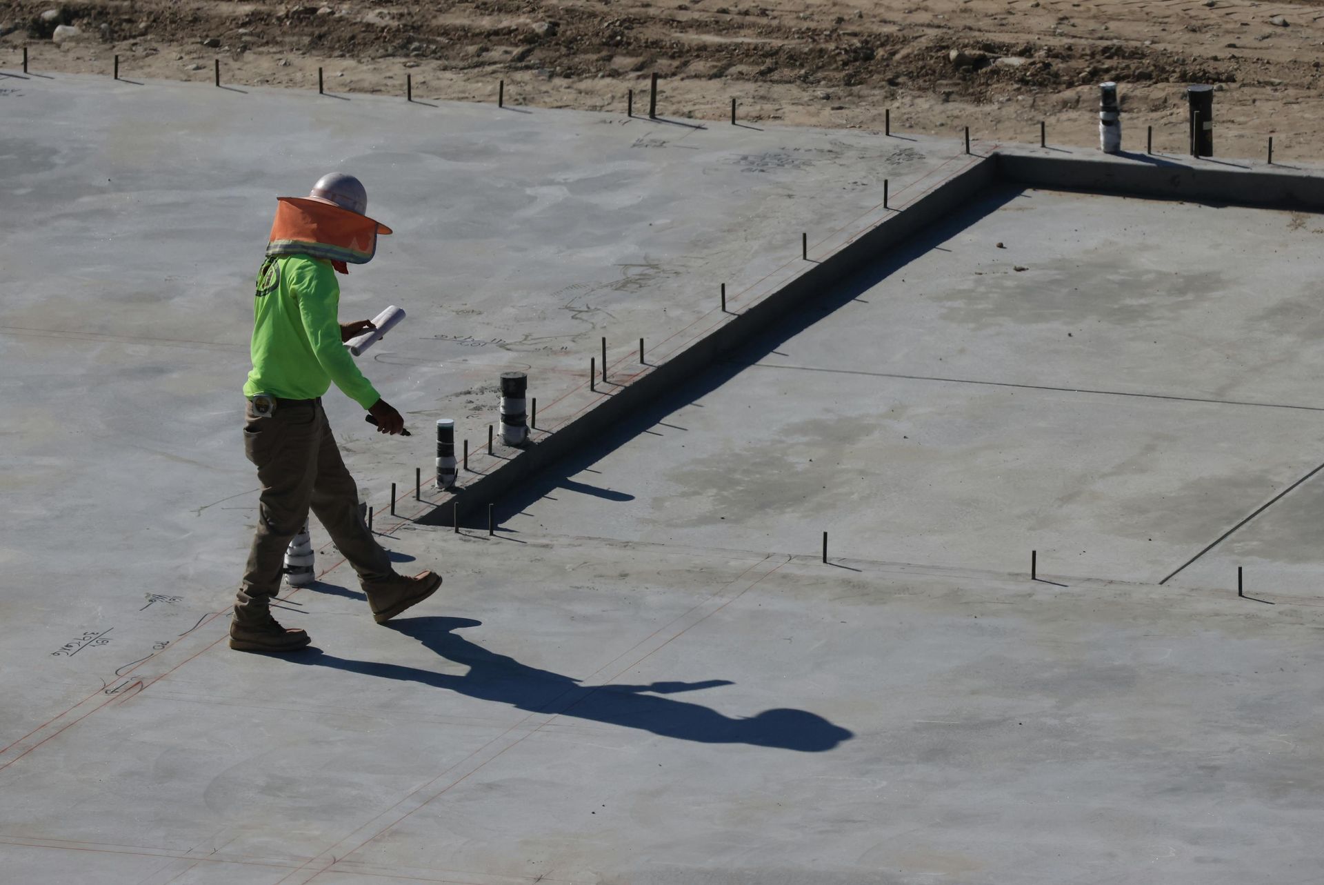 Worker in a neon vest and orange hard hat walking on a concrete rooftop near an angled wall.