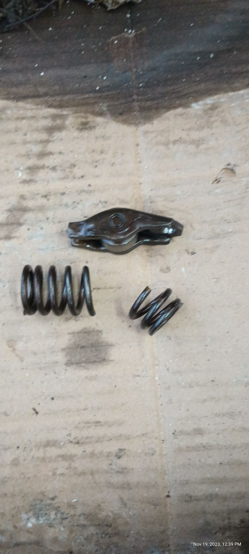 Engine rocker arm and two springs laid on a dirty, worn surface.