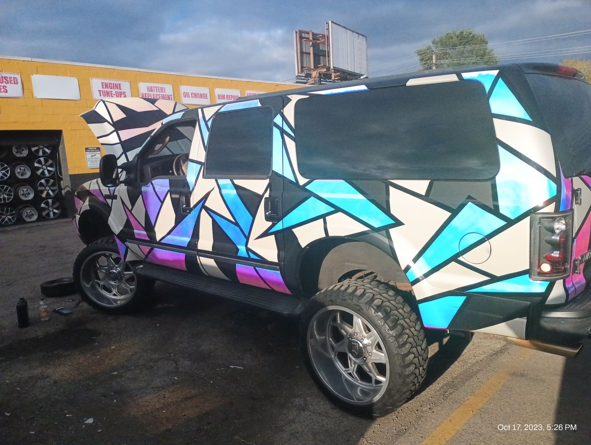 SUV with custom geometric wrap in white, blue, purple, and black; parked outside a tire shop.