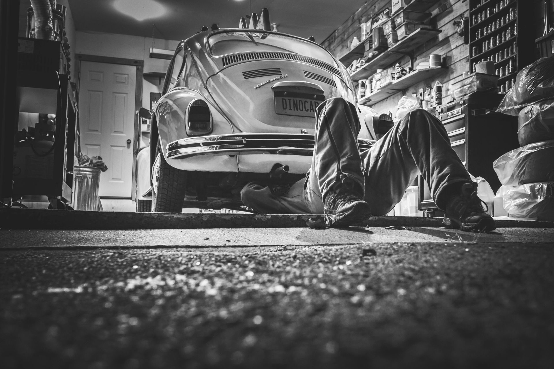 Man working on the underside of a classic car in a garage, black and white.