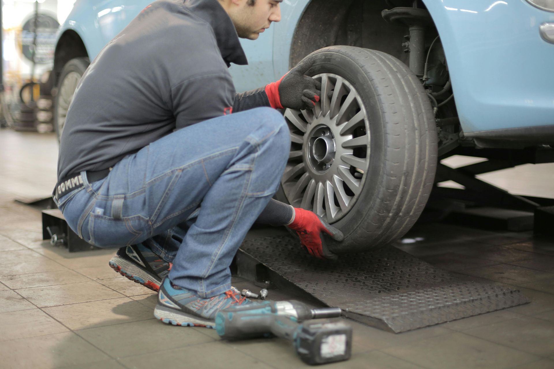 Mechanic in gray shirt and jeans changing a tire on a light blue car in a garage.