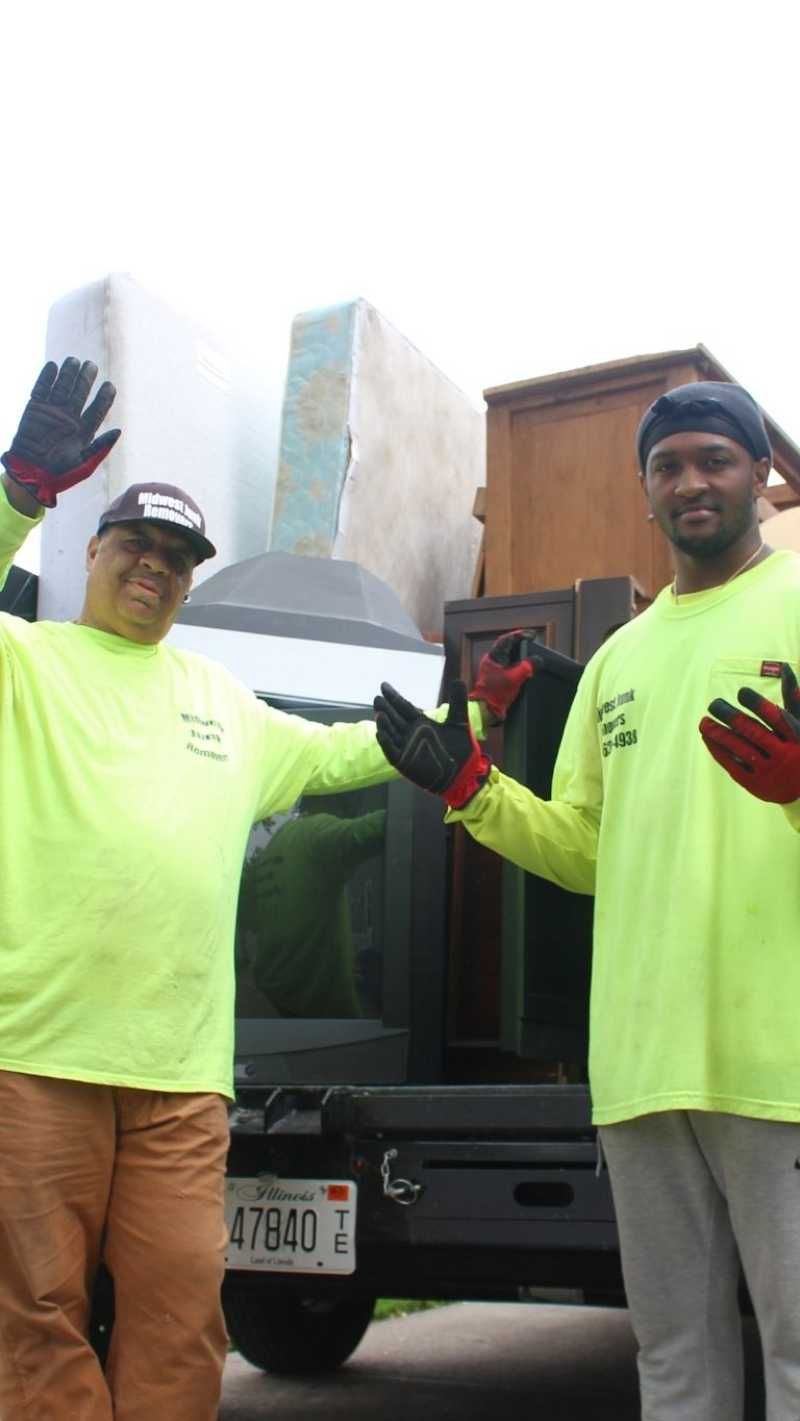 Two men in neon shirts wave next to a truck loaded with furniture and a mattress.