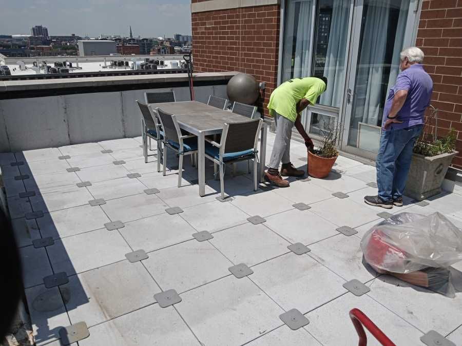 Two men on a rooftop patio, one tending a plant, the other watching. Brick building, table and chairs visible.