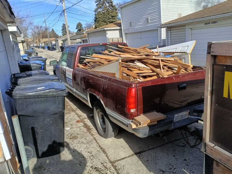 Red pickup truck filled with wood debris in an alley, next to trash bins, under a sunny sky.