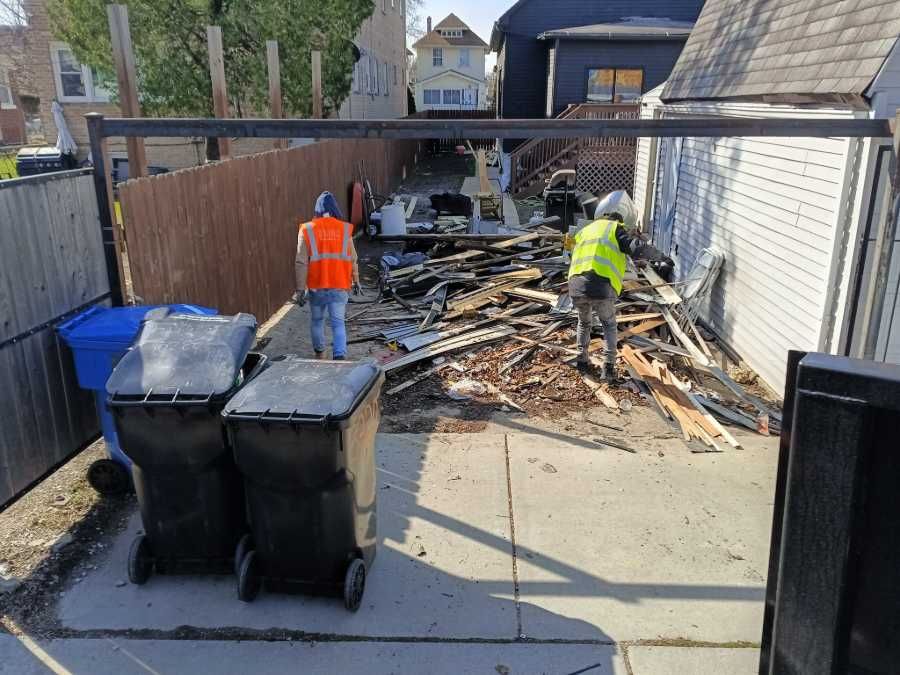 Two people in safety vests clearing debris in a backyard with garbage bins, fence, and houses.