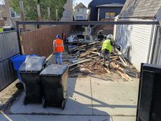 Two people in safety vests clearing debris in a backyard with garbage bins, fence, and houses.