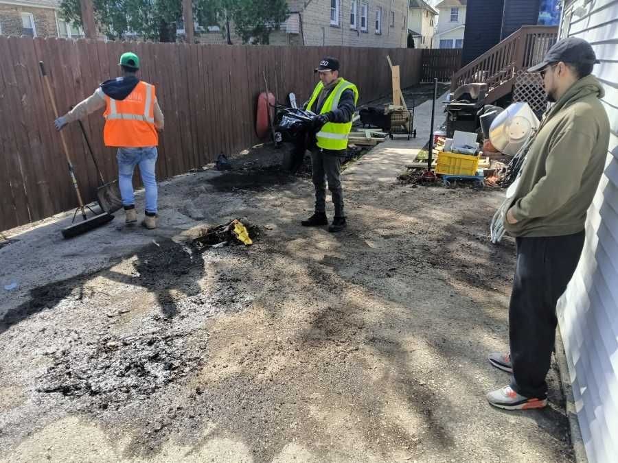 Three workers in safety vests are clearing a backyard. One rakes, another holds a tool, and one observes near a building.