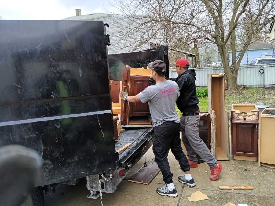 Two men loading furniture into a black dumpster truck in a yard. One wears a red cap.