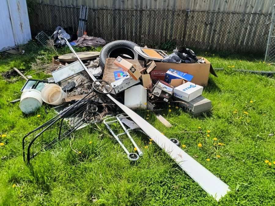 Pile of trash in a grassy yard: tire, boxes, metal, plastic, and debris sit near a fence.
