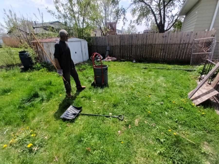Man stands in a grassy backyard, shovel on ground. Barrel fire pit, wooden fence, and shed in view.