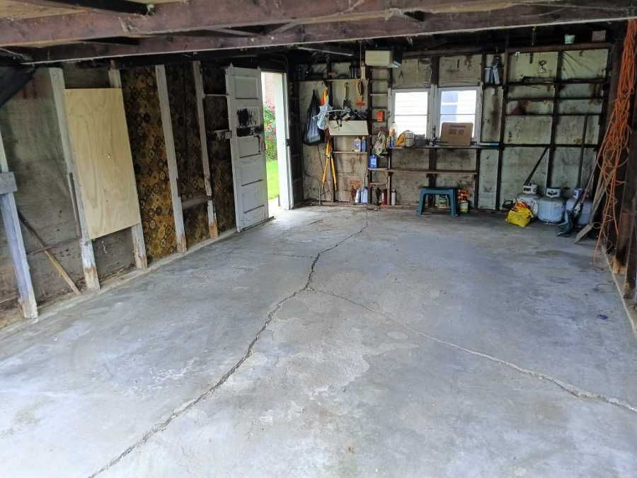 Bare-walled garage with cracked concrete floor, doorway, tools, and supplies visible.