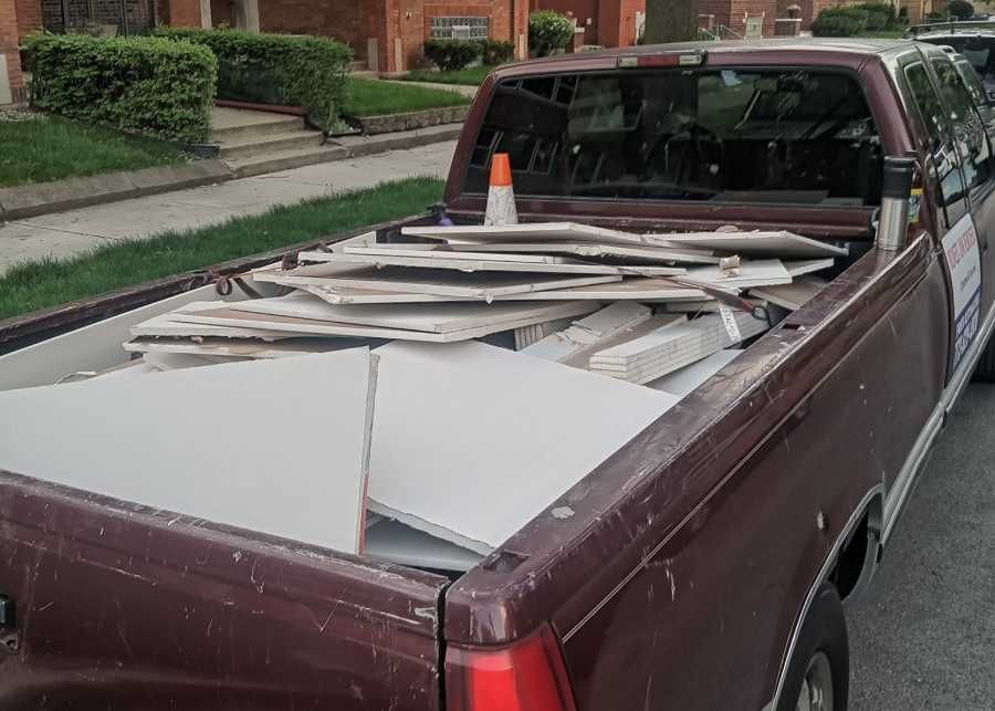 Red pickup truck bed filled with discarded white tiles and debris, parked on a street with a residential setting.