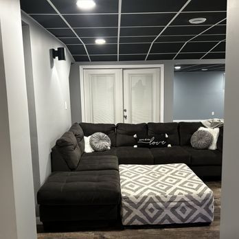 A living room with a black sectional couch and an ottoman.