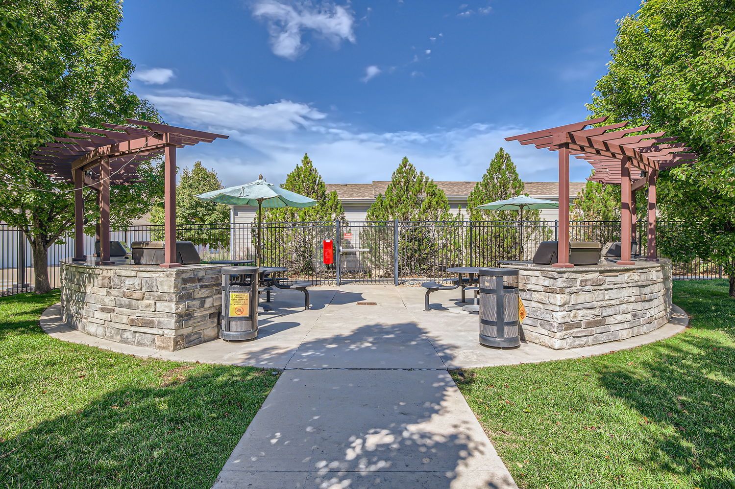 Outdoor community grilling area with stone planters, benches, umbrellas, and a paved walkway.
