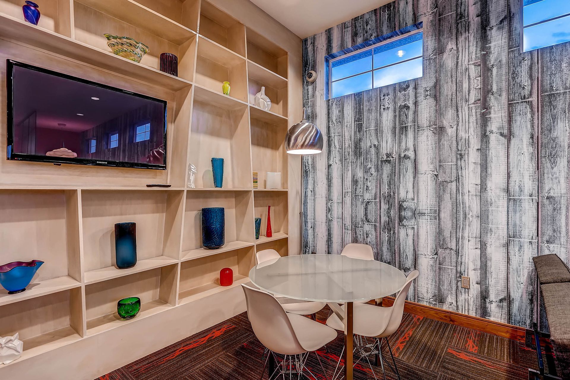 Interior community lounge with a round glass table, white chairs, and built-in shelves with decorative objects.