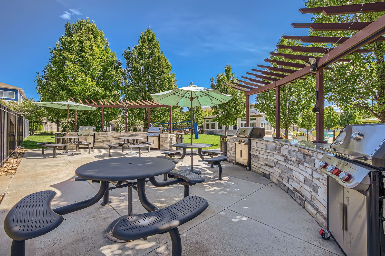 Outdoor community grilling area with gas grills, stone countertops, and umbrella-shaded tables near a pool.