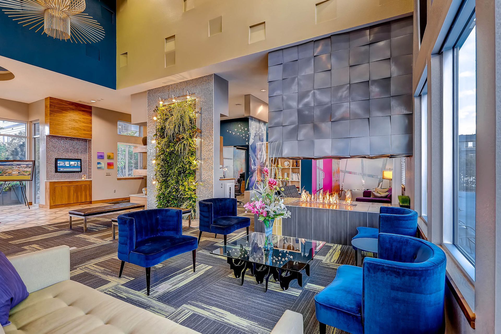 Modern residential lobby with blue velvet chairs, glass coffee table, a fireplace, and greenery.