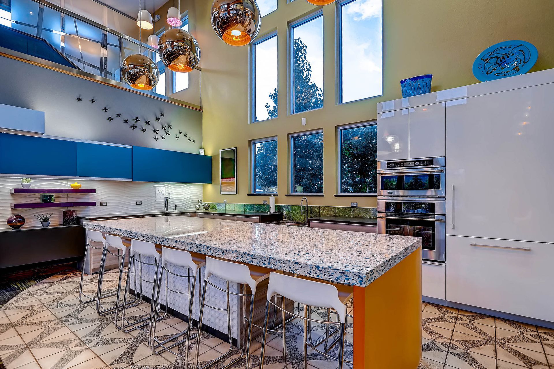 Modern kitchen with large island and tall windows; yellow, blue, and white color scheme.