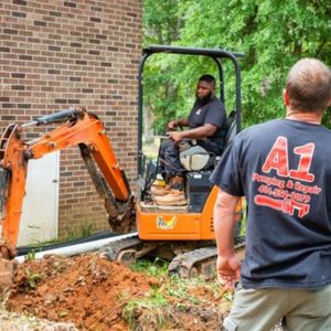 A man wearing a black a1 shirt stands in front of an excavator