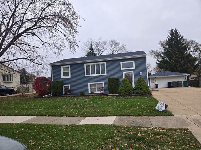 Two-story blue house with a green lawn on a cloudy day. Small shrubs and a garage are visible.
