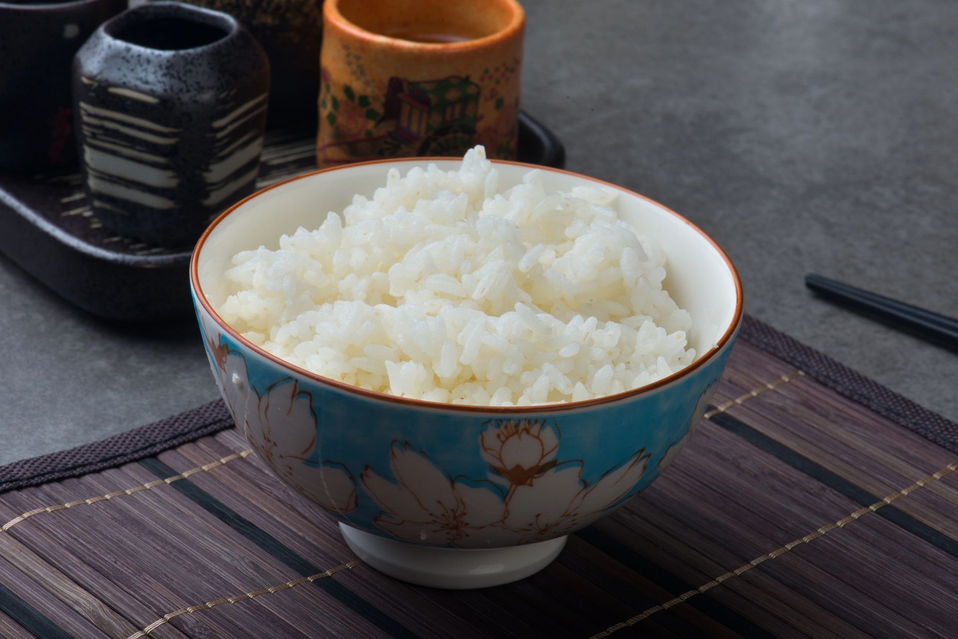Cuenco de arroz blanco sobre una estera de bambú, con un cuenco floral decorativo y tazas de té en el fondo.