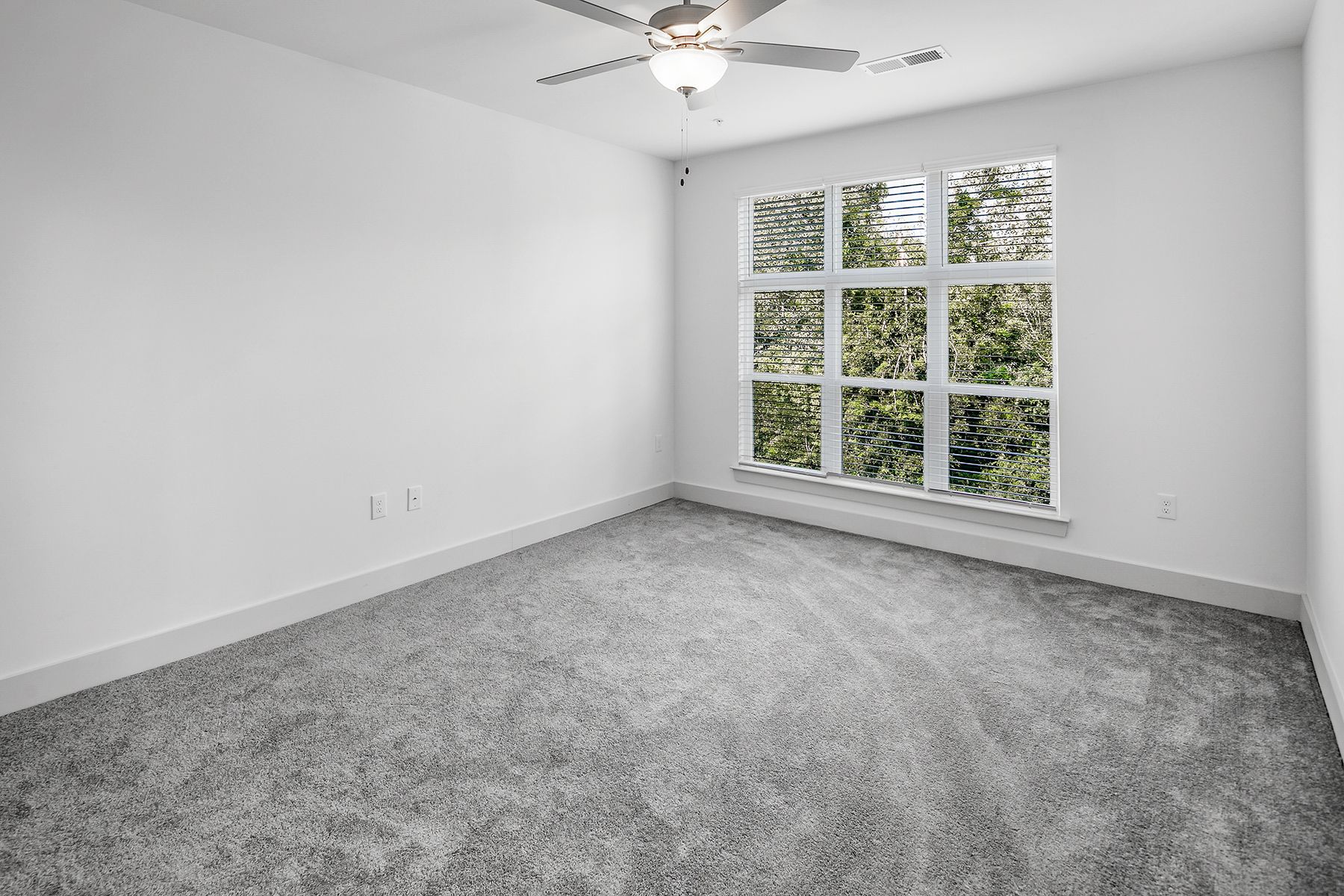 Empty room with gray carpet, white walls, large window with trees outside. Ceiling fan.