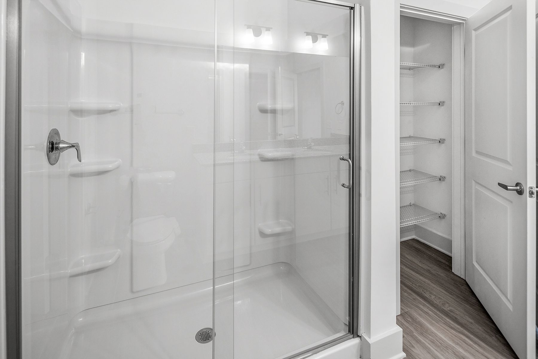 White shower with glass door, shelves, and a walk-in closet with glass shelves to the right.