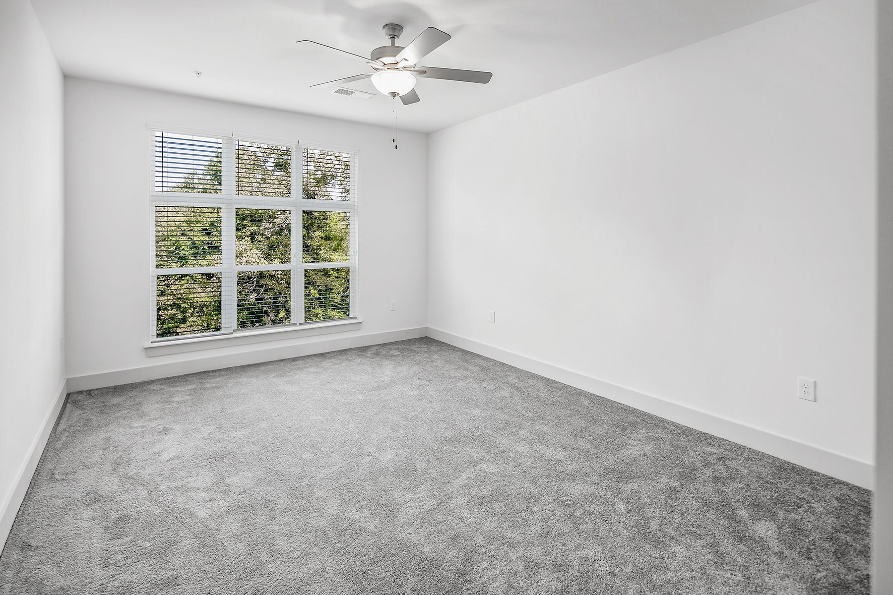 Empty bedroom with gray carpet, large window with trees, and a ceiling fan.