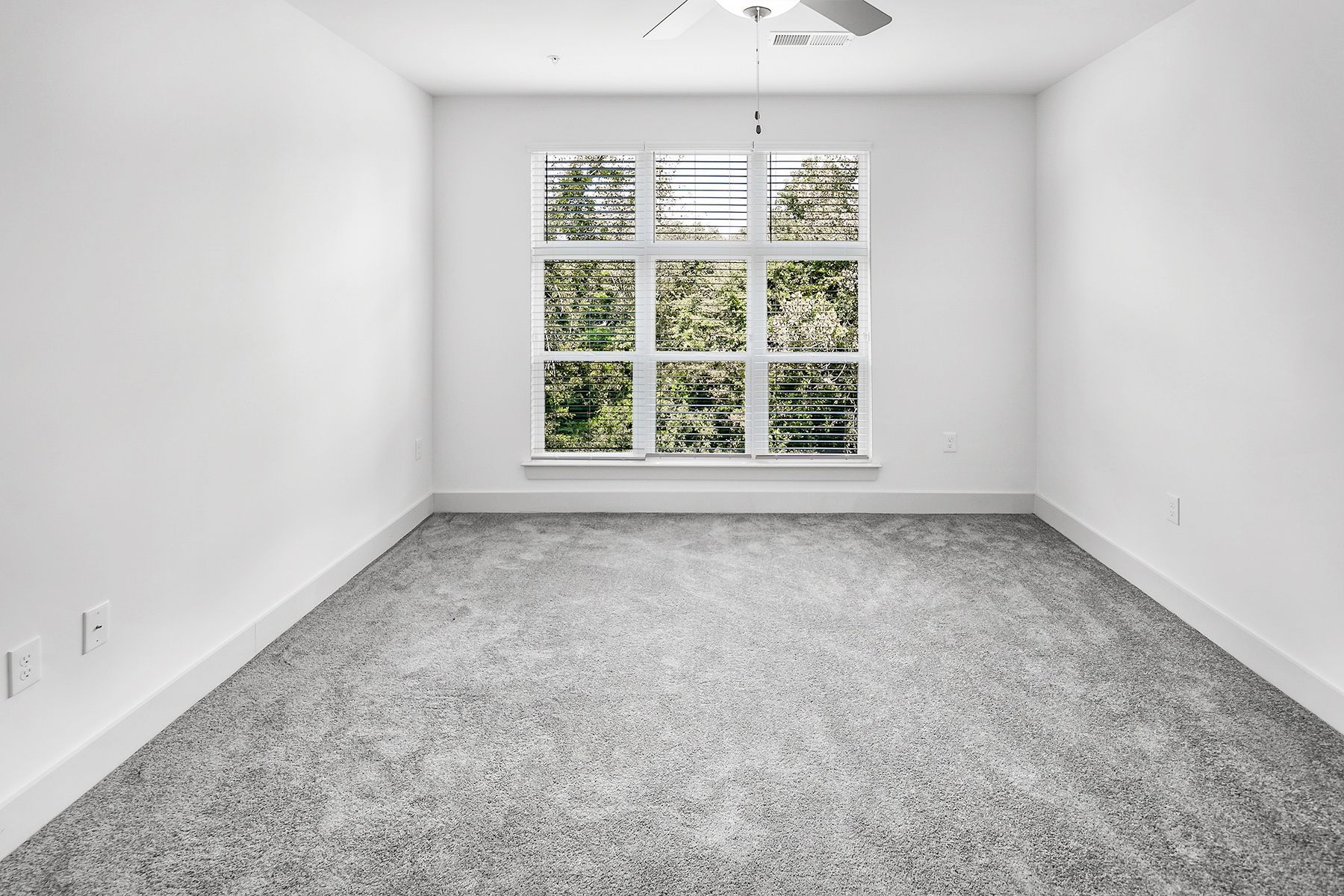 Empty room with gray carpet, white walls, and a large window overlooking green foliage.