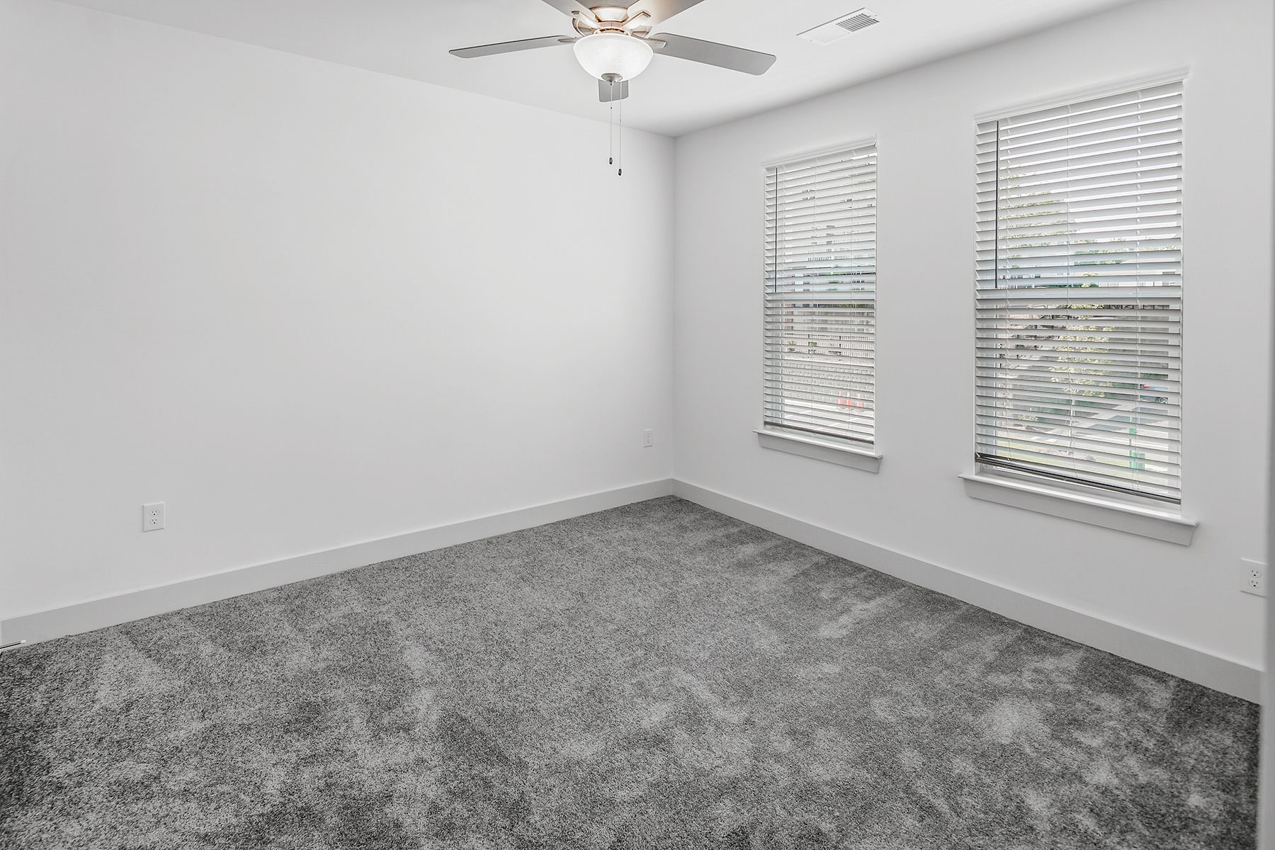 Empty bedroom with gray carpet, white walls, two windows with blinds, and a ceiling fan.