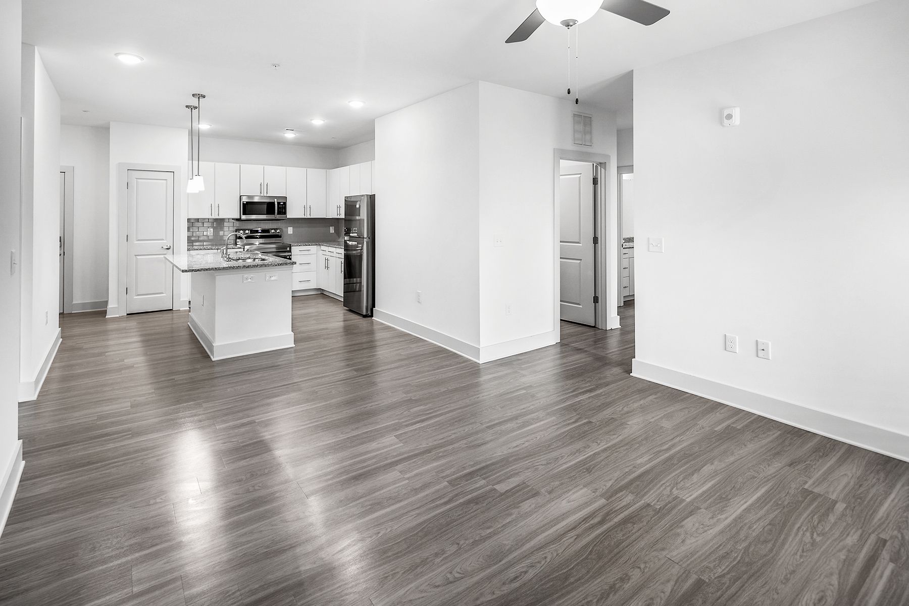Empty modern apartment interior with wood floors, white walls, kitchen, and ceiling fan.
