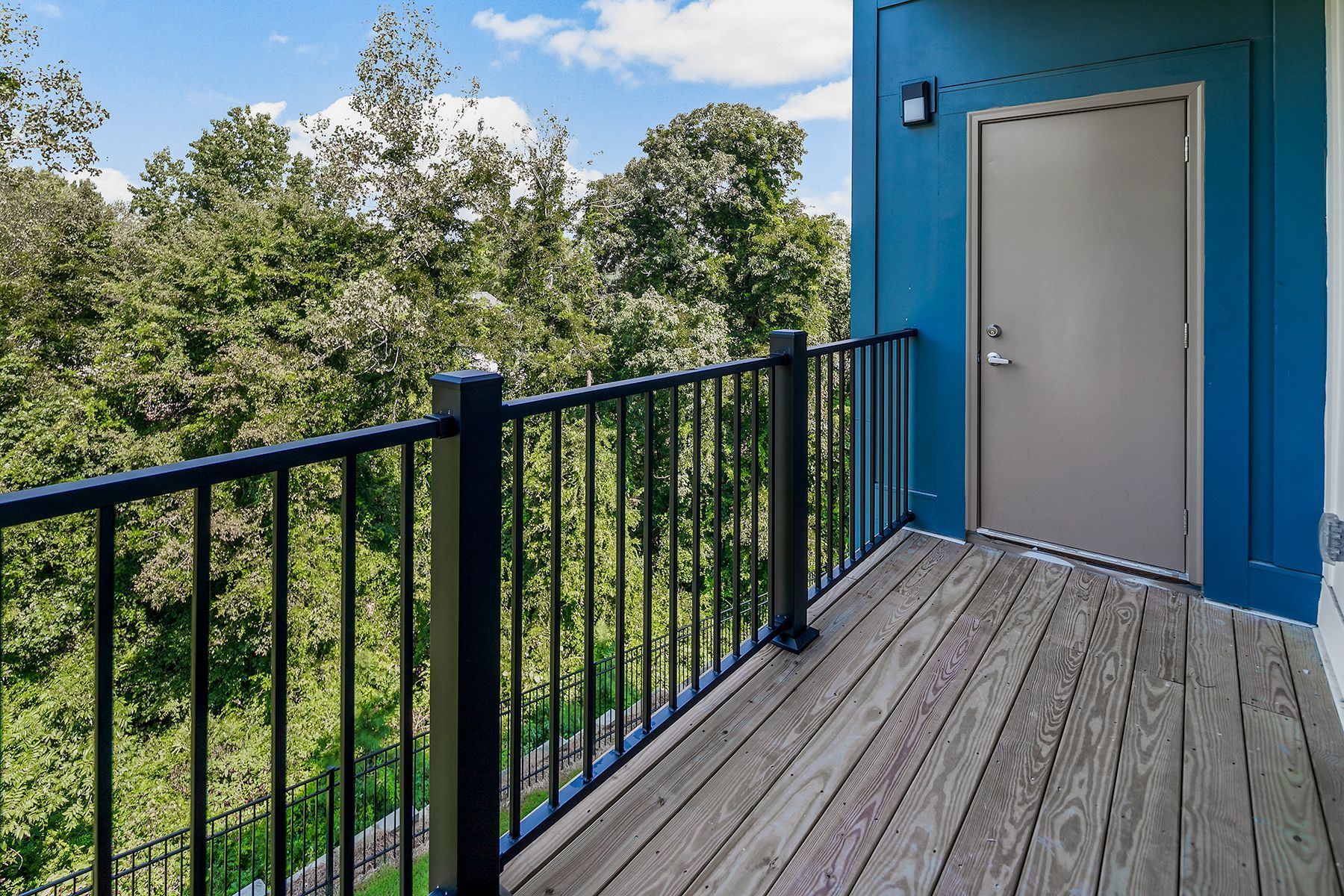 Balcony with black railing and wood floor, teal wall with a door, overlooking a green tree-filled landscape.