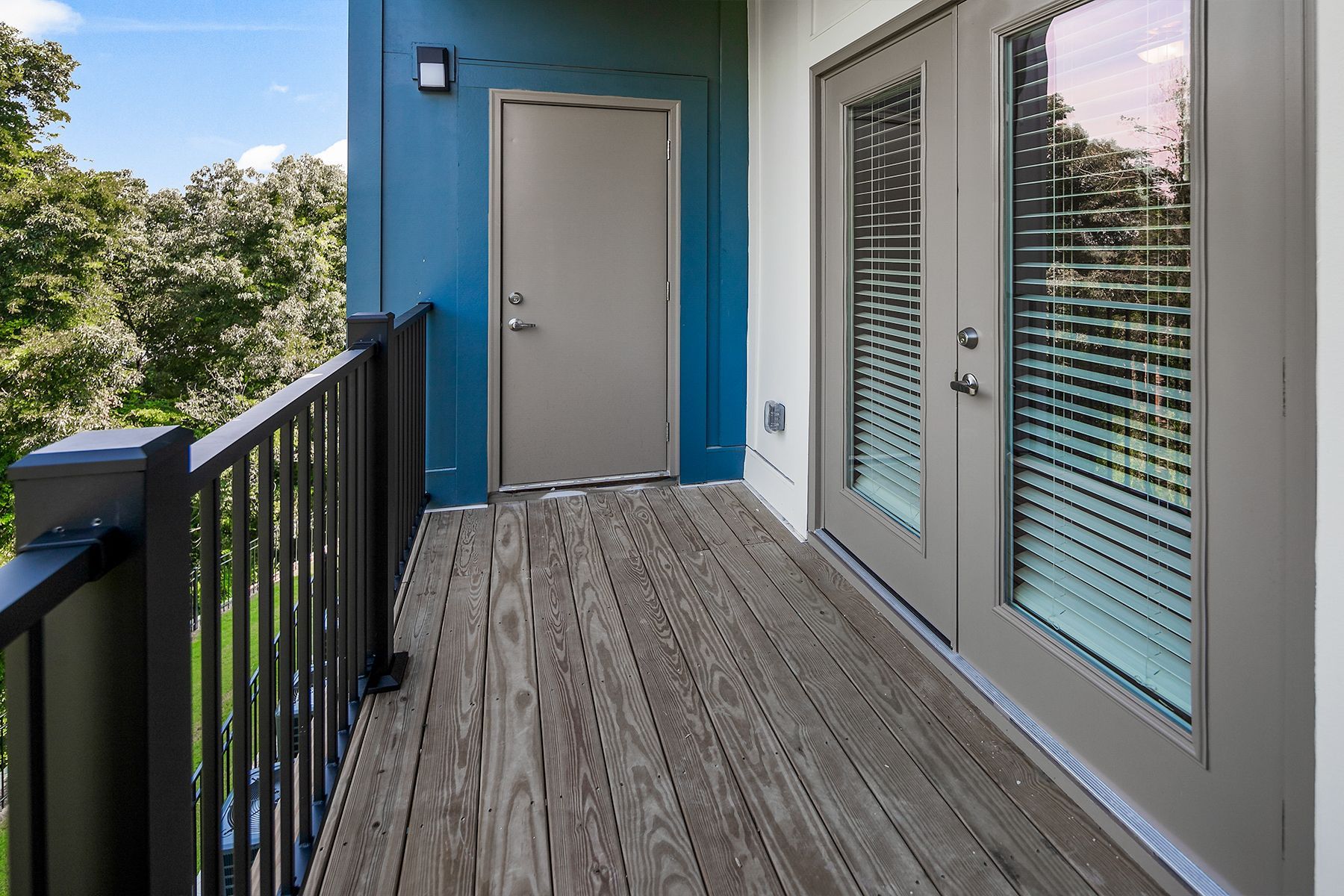 Balcony with gray wood planks, black railing, blue wall, and door, looking onto trees.