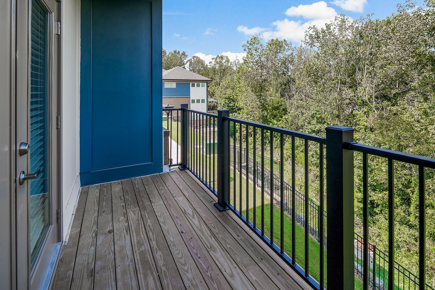 Balcony with wood decking and black railing, blue door, and view of trees and a building.