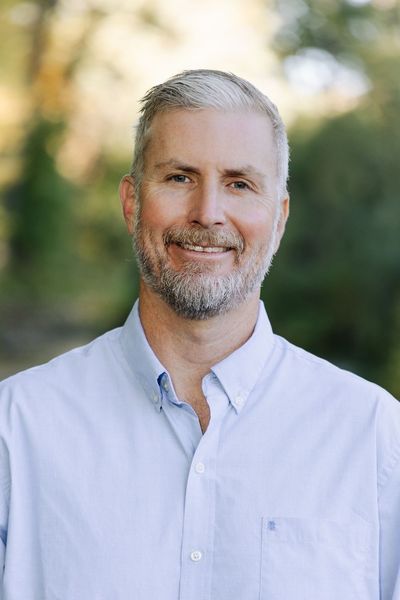 Man with graying hair and beard smiles in a light blue button-down shirt outdoors.