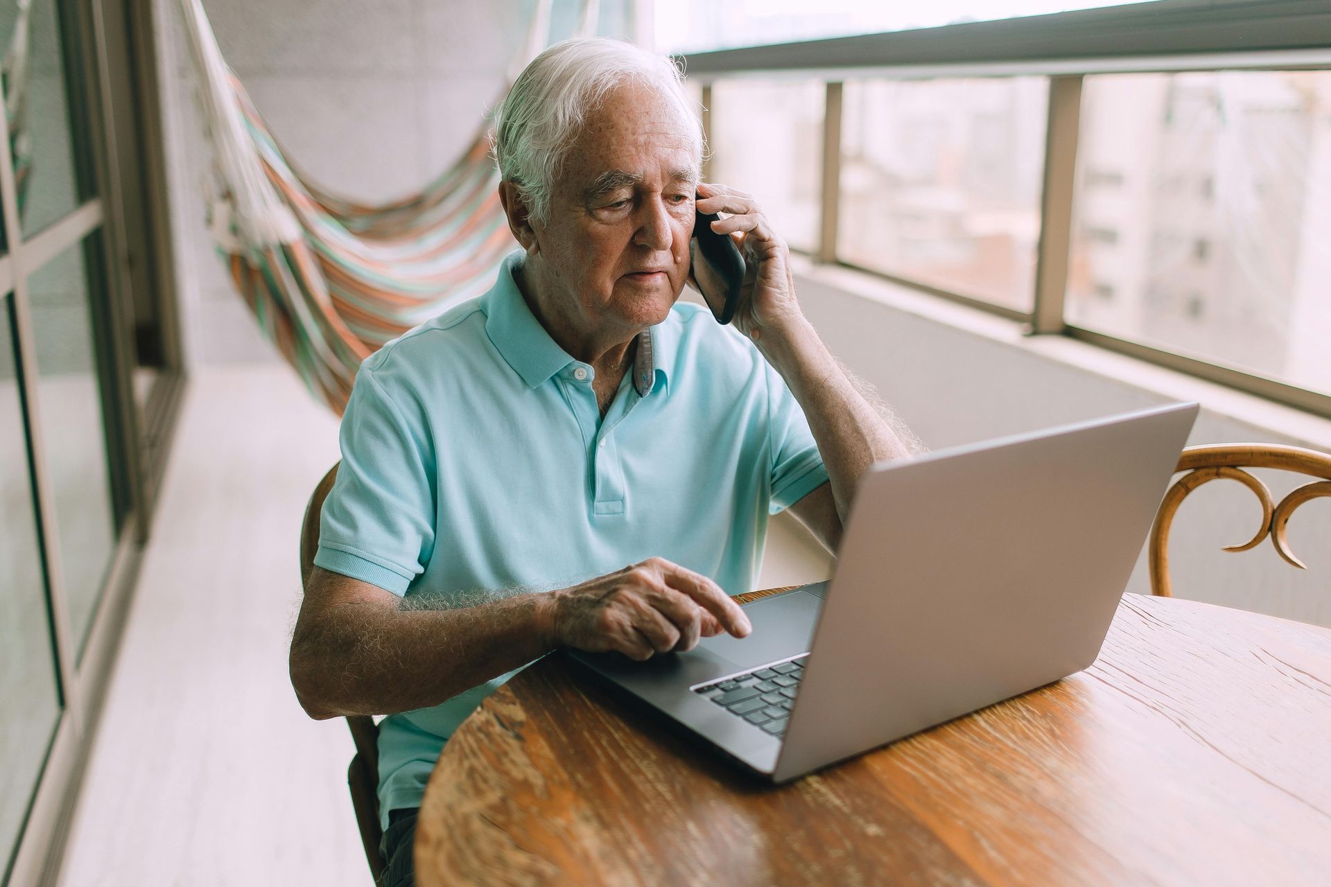 An elderly man is sitting at a table using a laptop and talking on a cell phone.
