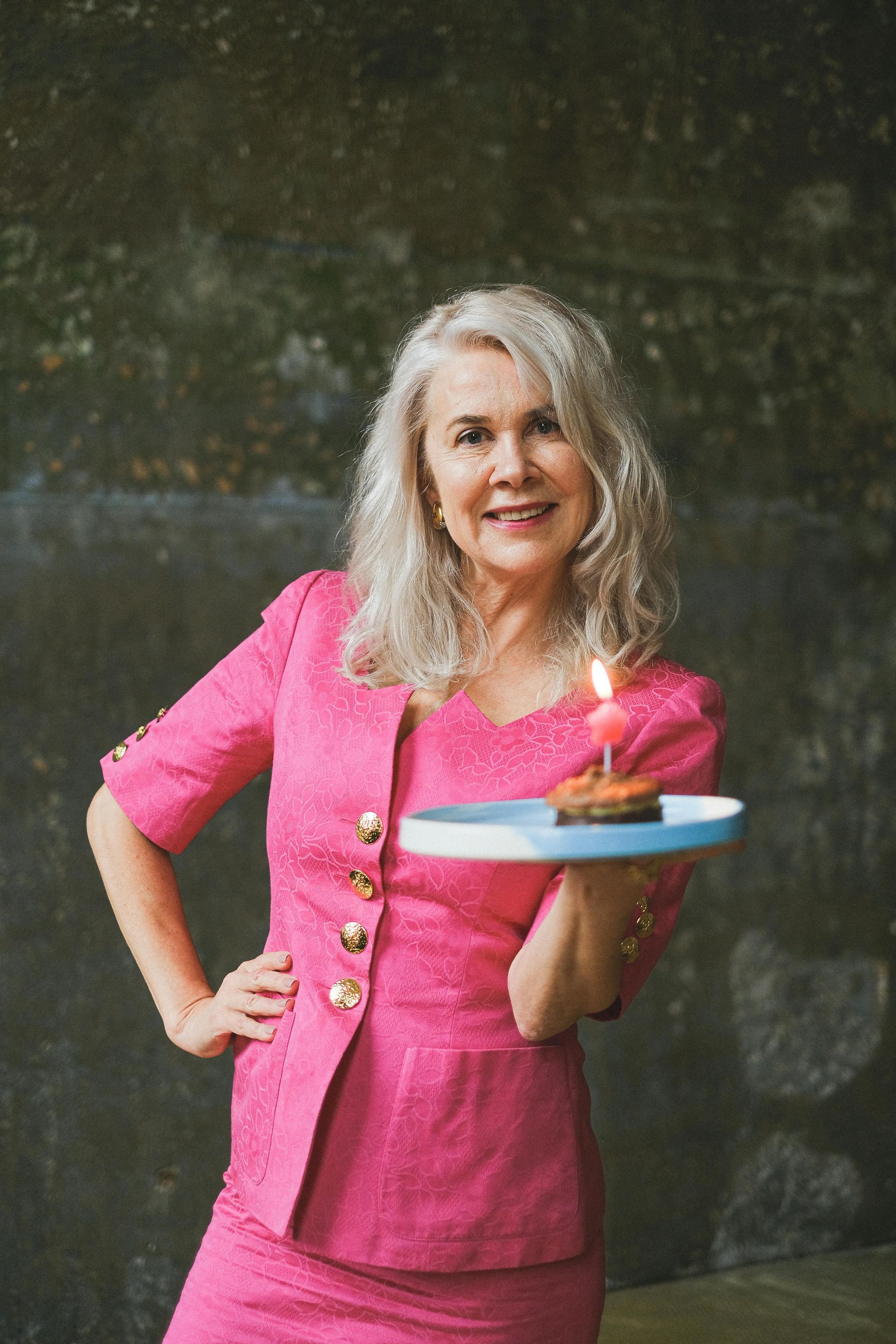 A mature woman in a pink suit is holding a plate with a cake on it.