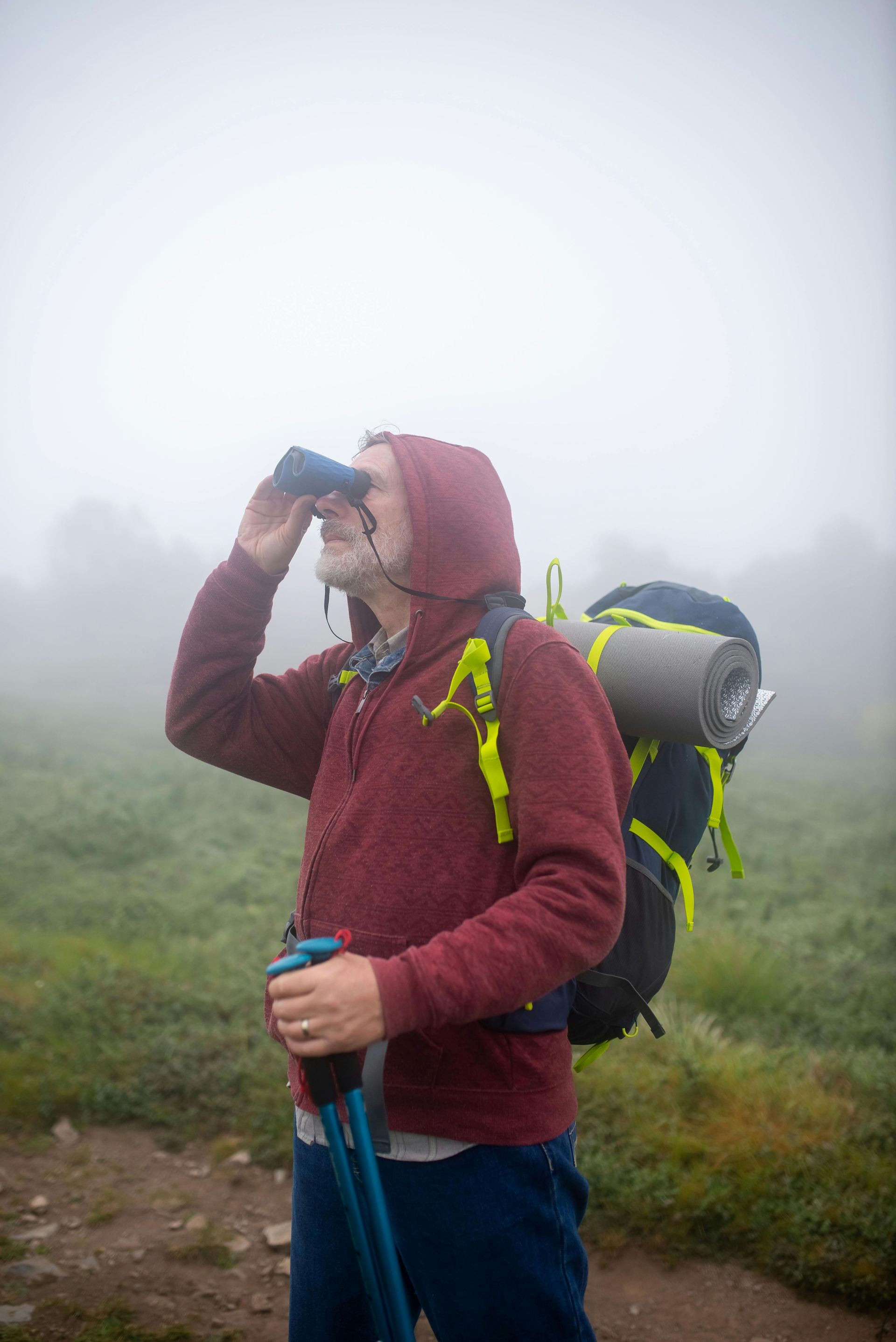 A man with a white beard and wearing a backpack is looking through binoculars in the fog.