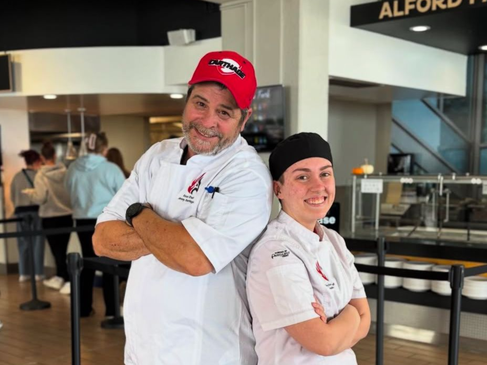 Two chefs pose in a cafeteria with arms crossed; one in a red hat, the other in a black hat.