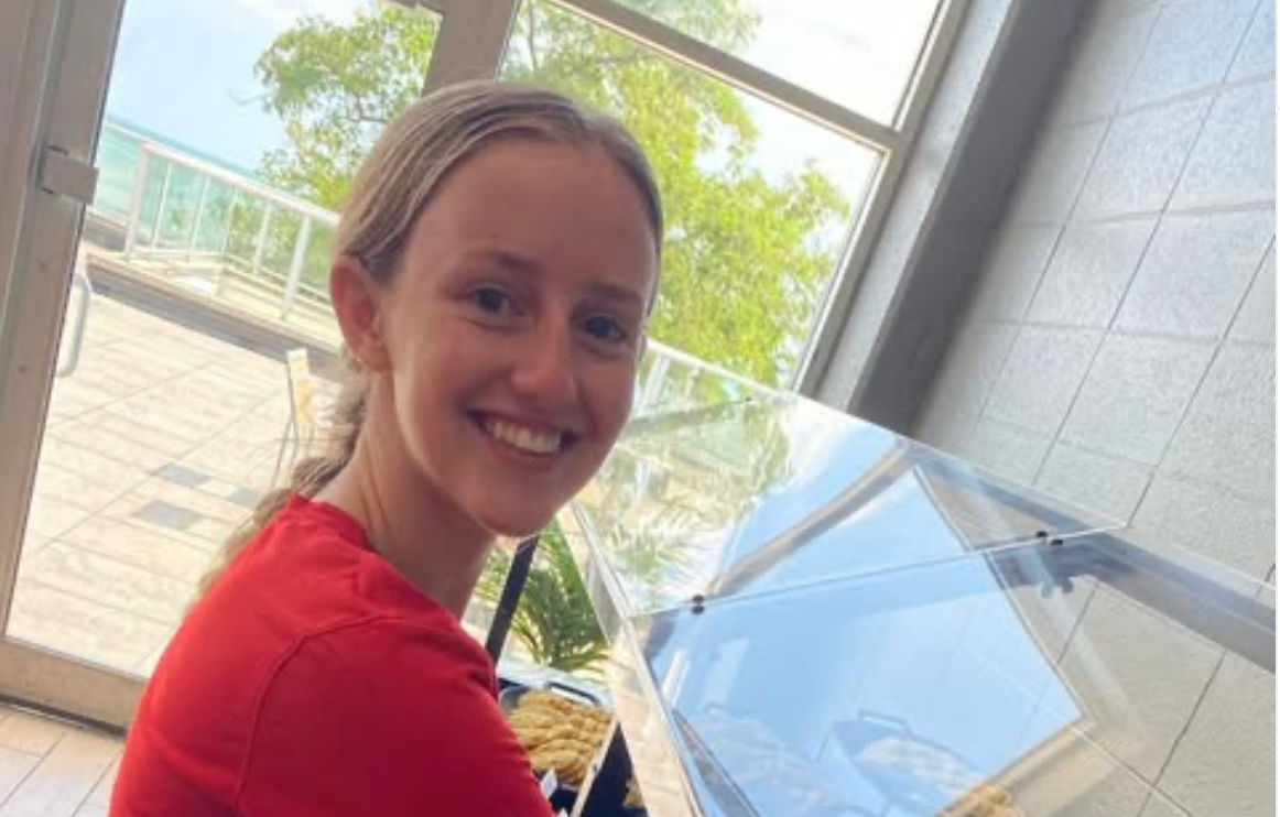 A smiling person in a red shirt stands indoors near a glass display case with a balcony.