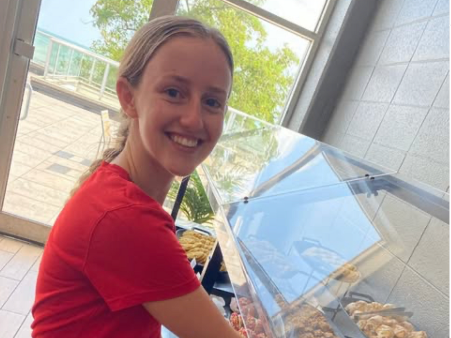 Woman in red shirt smiles at a clear food display case near a window.