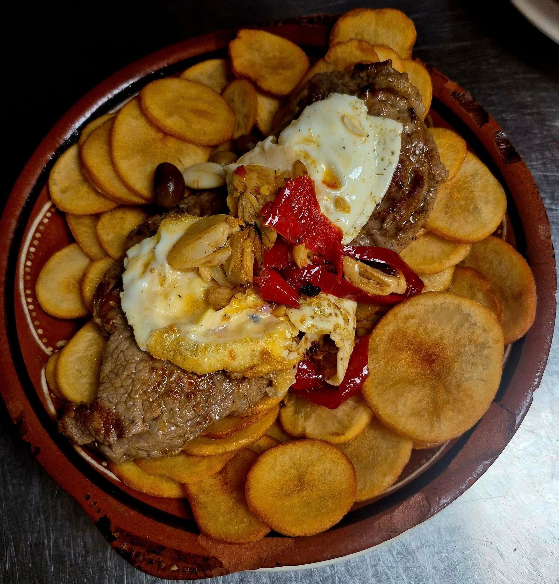 Steak with fried eggs, roasted peppers, and potato chips on a rustic brown plate.