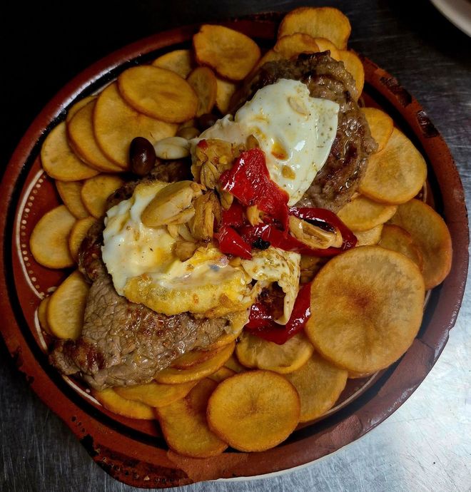 Steak and fried egg dish with potato chips, red peppers, and olive on a round, rustic plate.