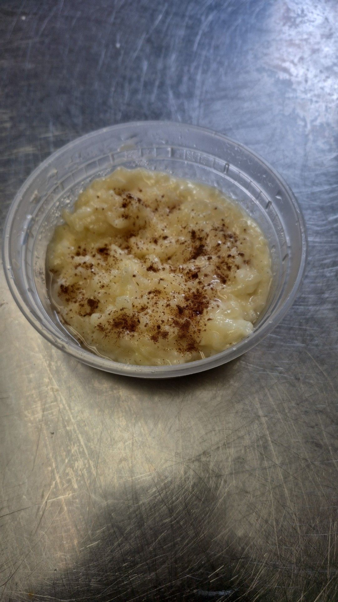 A small clear plastic container with cooked oatmeal sprinkled with cinnamon on a stainless steel surface.