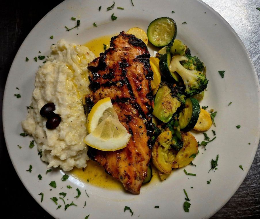 Grilled fish with lemon, mashed potatoes, roasted vegetables, and olives on a white plate.