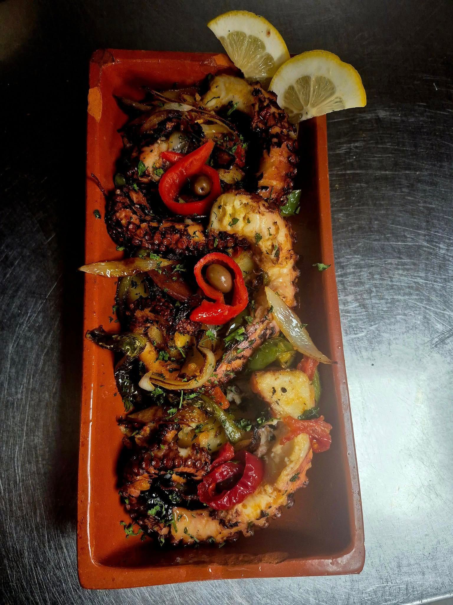 Grilled octopus with vegetables and lemon garnish on a brown rectangular plate.