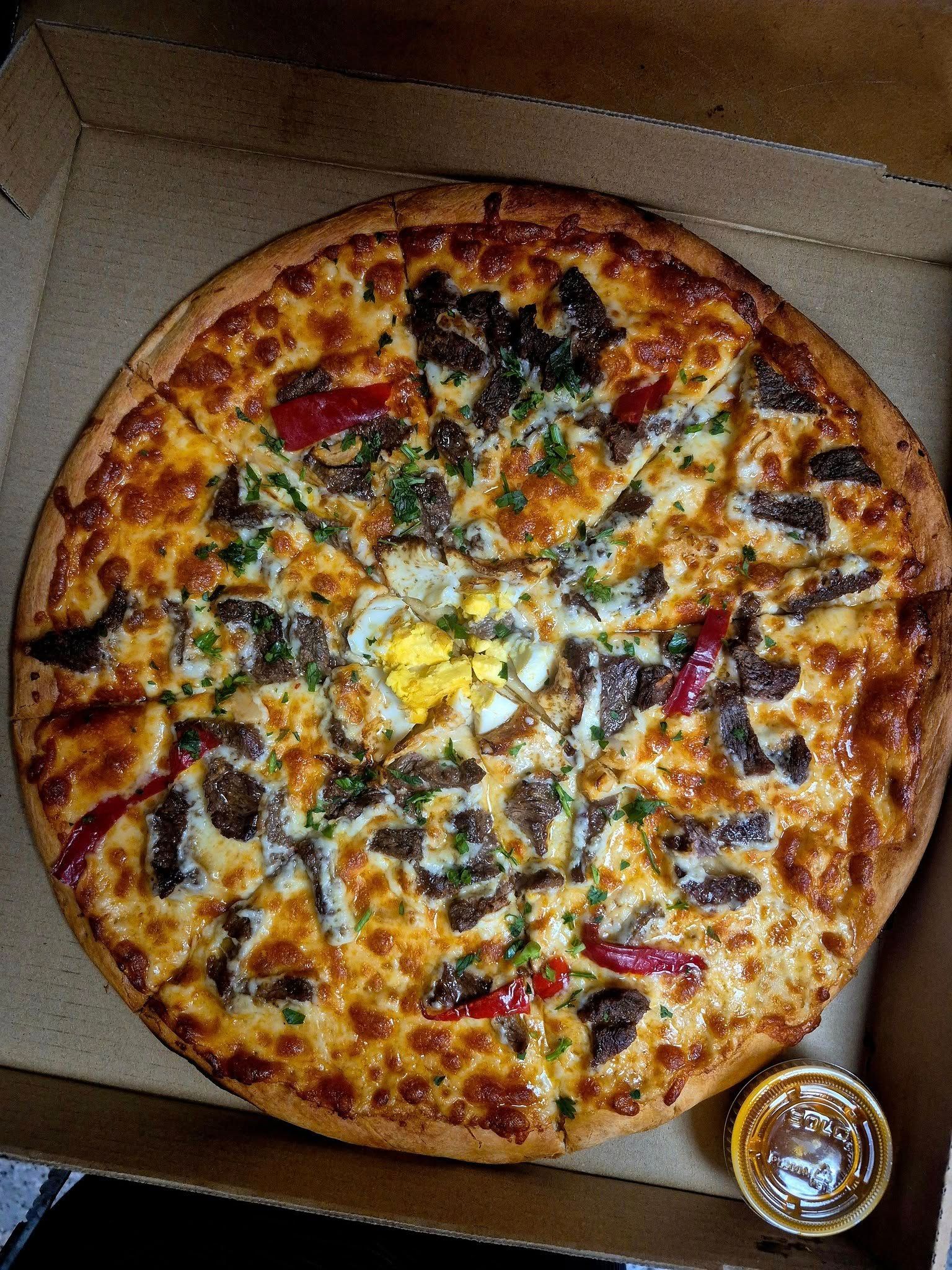 Pizza with toppings in a cardboard box, with a sauce container to the side.