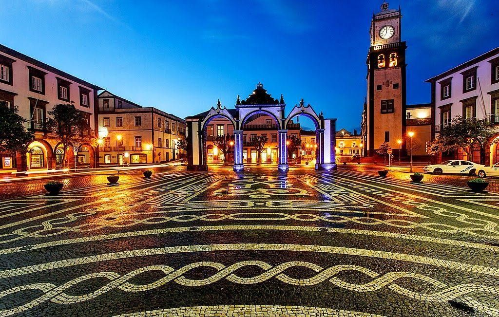 Night view of a cobblestone plaza with lit buildings and an archway, reflecting lights, in front of a clock tower.