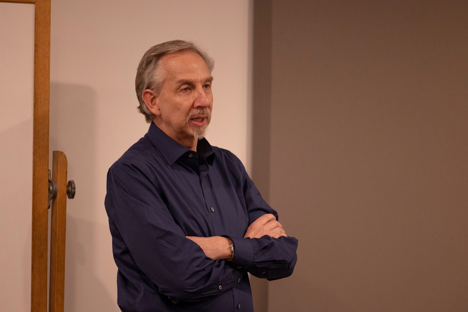A man in a blue shirt is standing with his arms crossed in front of a white board.